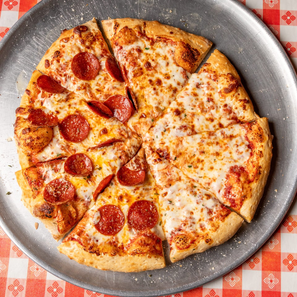 A pepperoni and cheese pizza, with some slices removed, on a metal tray over a red and white checkered tablecloth.