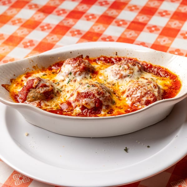 Baked meatballs in a white dish, covered in melted cheese and red sauce, on a checkered tablecloth.