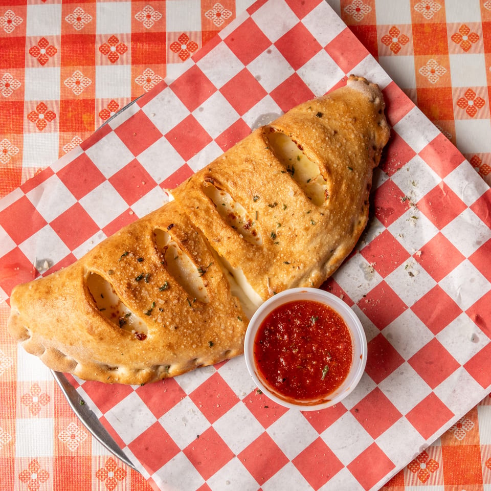 Golden-brown calzone with visible cheese filling and a side of marinara sauce on a checkered paper.