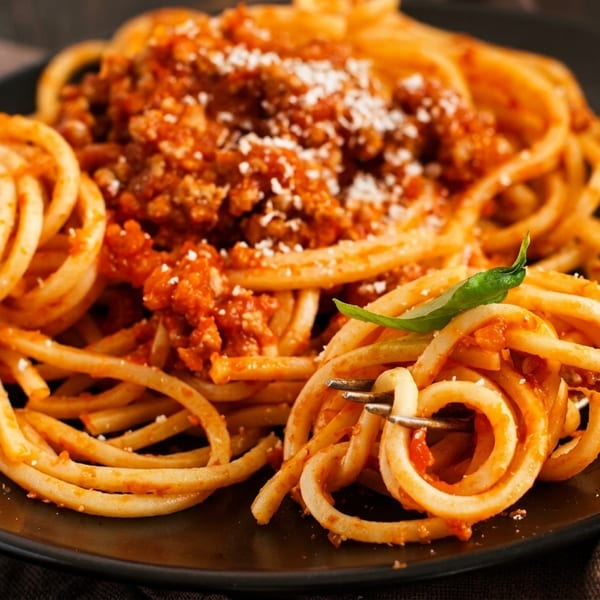 Close-up of bucatini pasta with bolognese sauce, parmesan, and basil, with a fork twirling noodles.