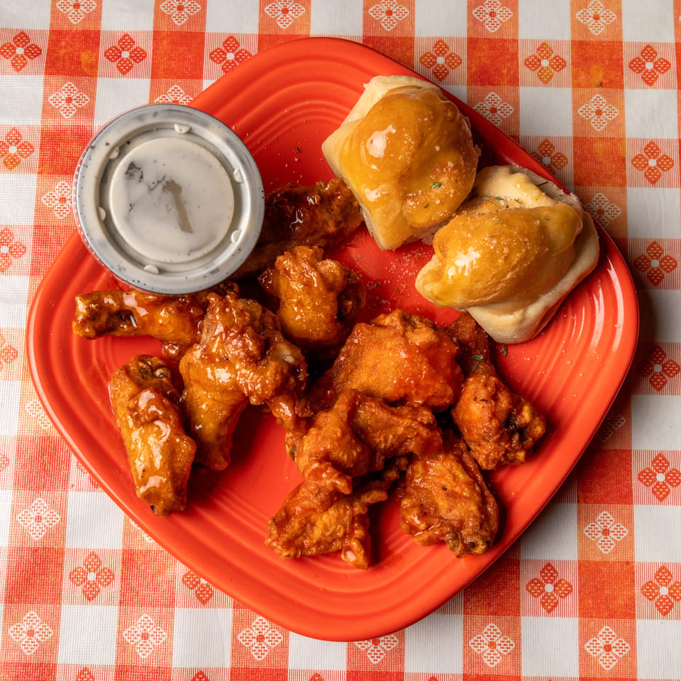 Orange plate of buffalo wings, garlic knots, and ranch dressing on a red and white checkered tablecloth.