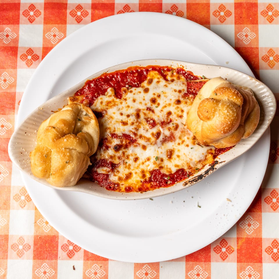 Baked pasta with melted cheese and tomato sauce, served with two garlic knots on a checkered tablecloth.